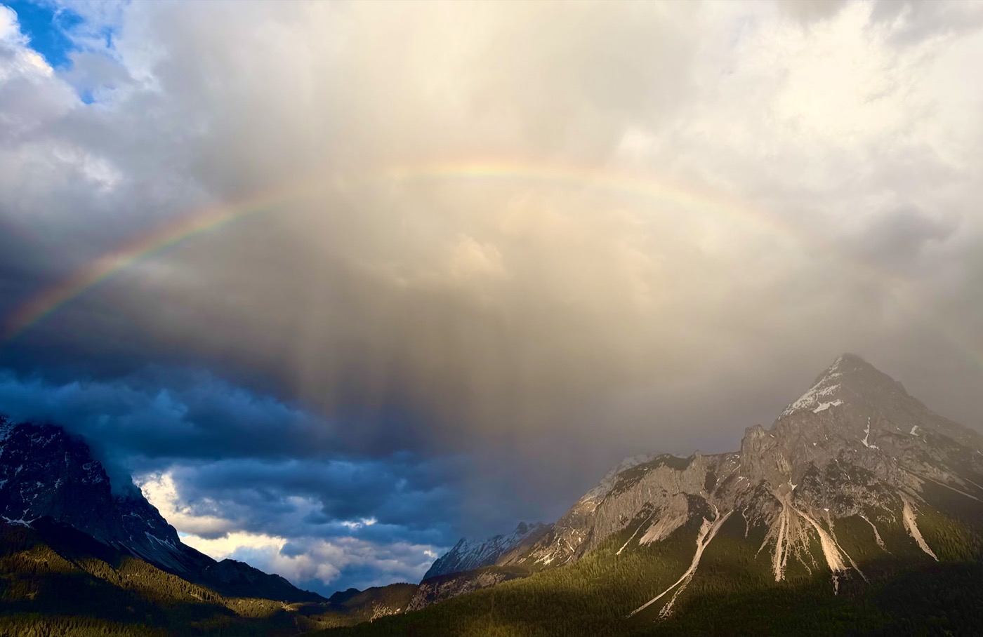 Regenbogen über einer Bergkette, während die Sonne durch die Regenwolken bricht